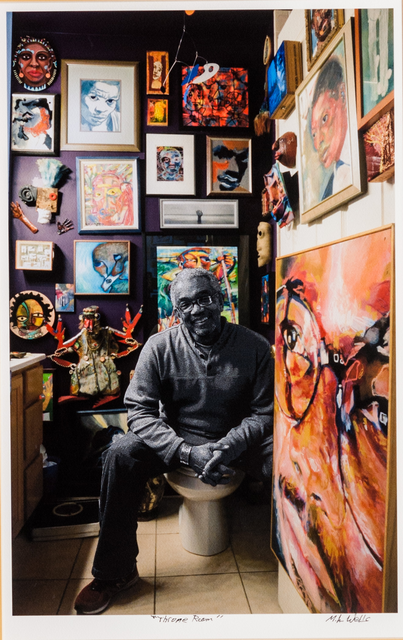 Photograph of Patric McCoy sitting down in a small room with the walls around him filled by art