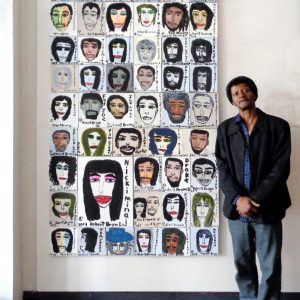 Photograph of Bobbie Brown standing in front of a wall with many of his portraits