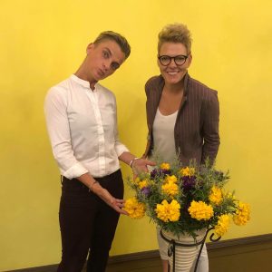 Photograph of Inga and Evija posing with yellow flowers in front of a yellow background