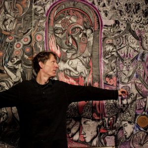 Photograph of Justin Duerr with his arms outstretched, looking over his shoulder at his large-scale drawing behind him