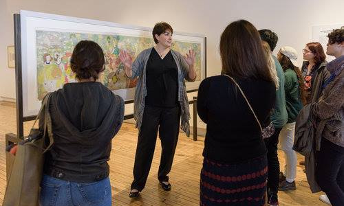 Photograph of Leisa Rundquist standing in front of a panoramic piece by Henry Darger with guests around her