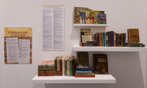 Photograph of books from Henry Darger's collection on three selves with exhibition signage in the top left corner