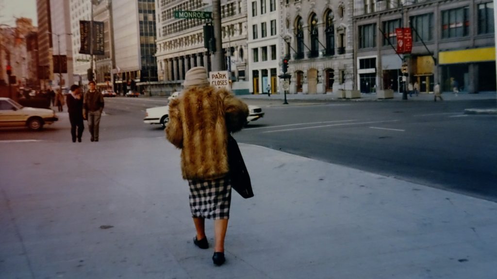 Film still of Lee Godie wearing a brown fur coat, walking away from the camera