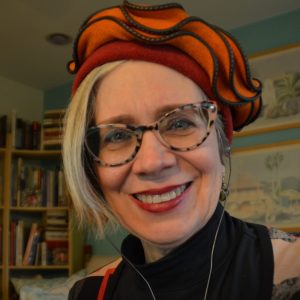 Photograph of a woman identified as Deb Kerr smiling and wearing a red-orange hat, colorful eyeglasses and a black turtleneck sweater