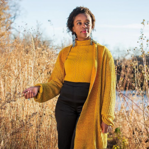 Photograph of a person, identified as Abena Motaboli, wearing a yellow sweater standing in a field of tall grasses with water in the background