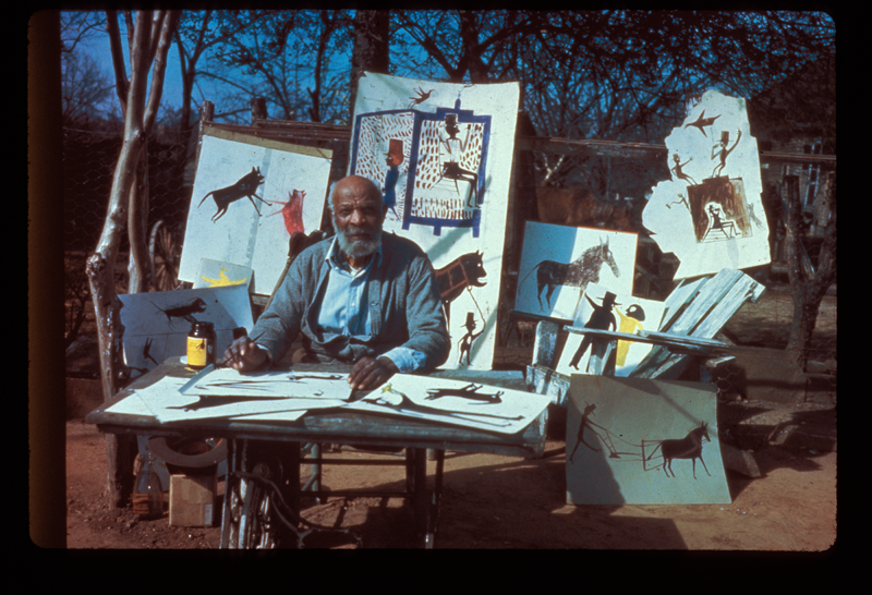 Photograph of artist Bill Traylor, sitting outside at a table with drawings and paintings around him