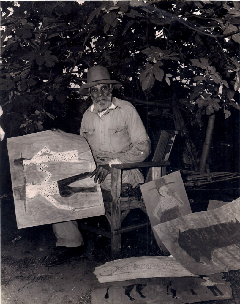 Photograph of artist Bill Traylor, sitting outside in front of trees with an artwork in his hands