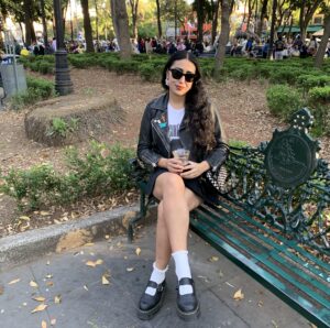 Photo of a Latina, identified as Mayra Palafox, sitting on a park bench, wearing sunglasses 