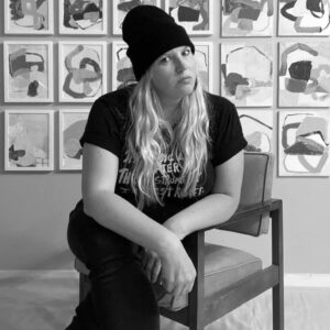 Black and white photo of a white woman, identified as Melissa Smith, sitting on a chair in front of a wall filled with art
