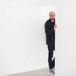 Photo of a man, identified as Jan Tichy, wearing all black, emerging from a doorway in an all-white room