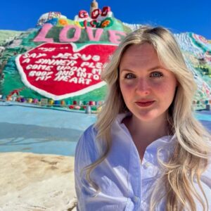 Photo of a white woman, identified as Annalise Flynn, wearing a white shirt, standing in front of Salvation Mountain