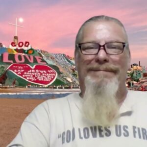 Photo of a man, identified as Bob Levesque, smiling, wearing eye glasses, with Salvation Mountain in the background